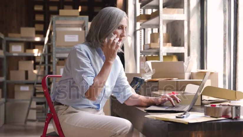 stock-footage-older-female-dropshipping-business-owner-using-laptop-talking-on-phone-in-warehouse-mature-woman